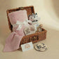 Wicker basket with baby items including a pink blanket, stuffed toy, and heart-shaped card on a beige background.