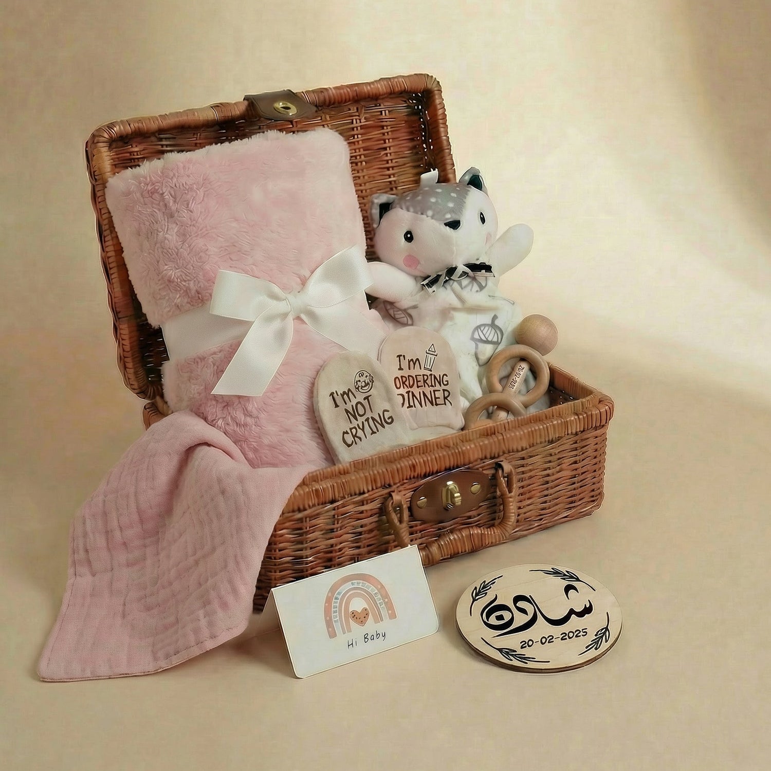 Wicker basket with baby items including a pink blanket, stuffed toy, and heart-shaped card on a beige background.